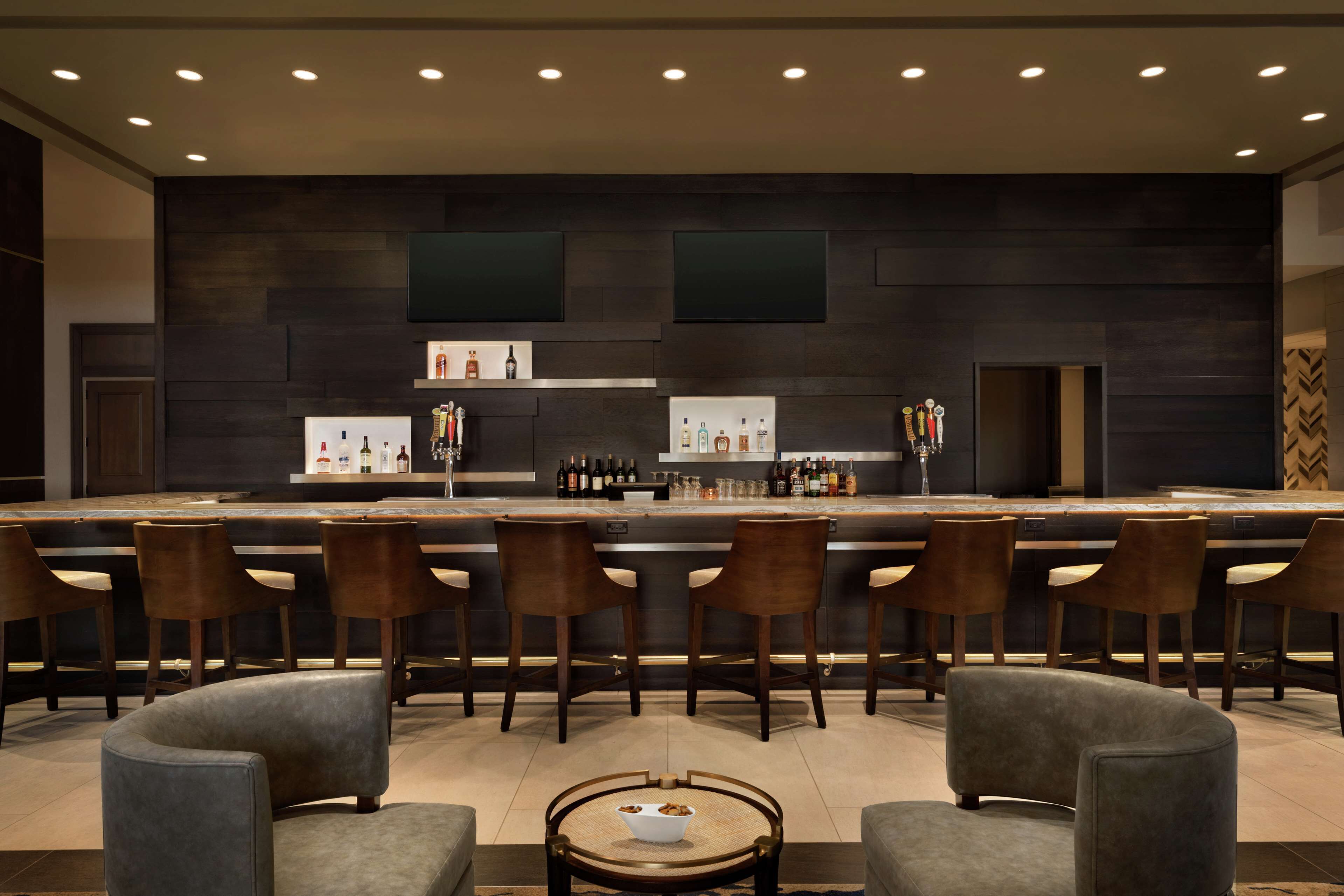 A modern bar area with a long counter, multiple bar stools, and a seating area with sofas and a coffee table.