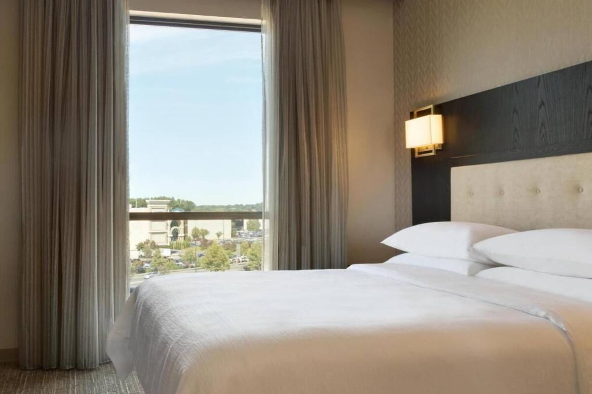 Une chambre d’hôtel avec un grand lit, une tête de lit dotée d’un luminaire et une vue sur la ville par la fenêtre.