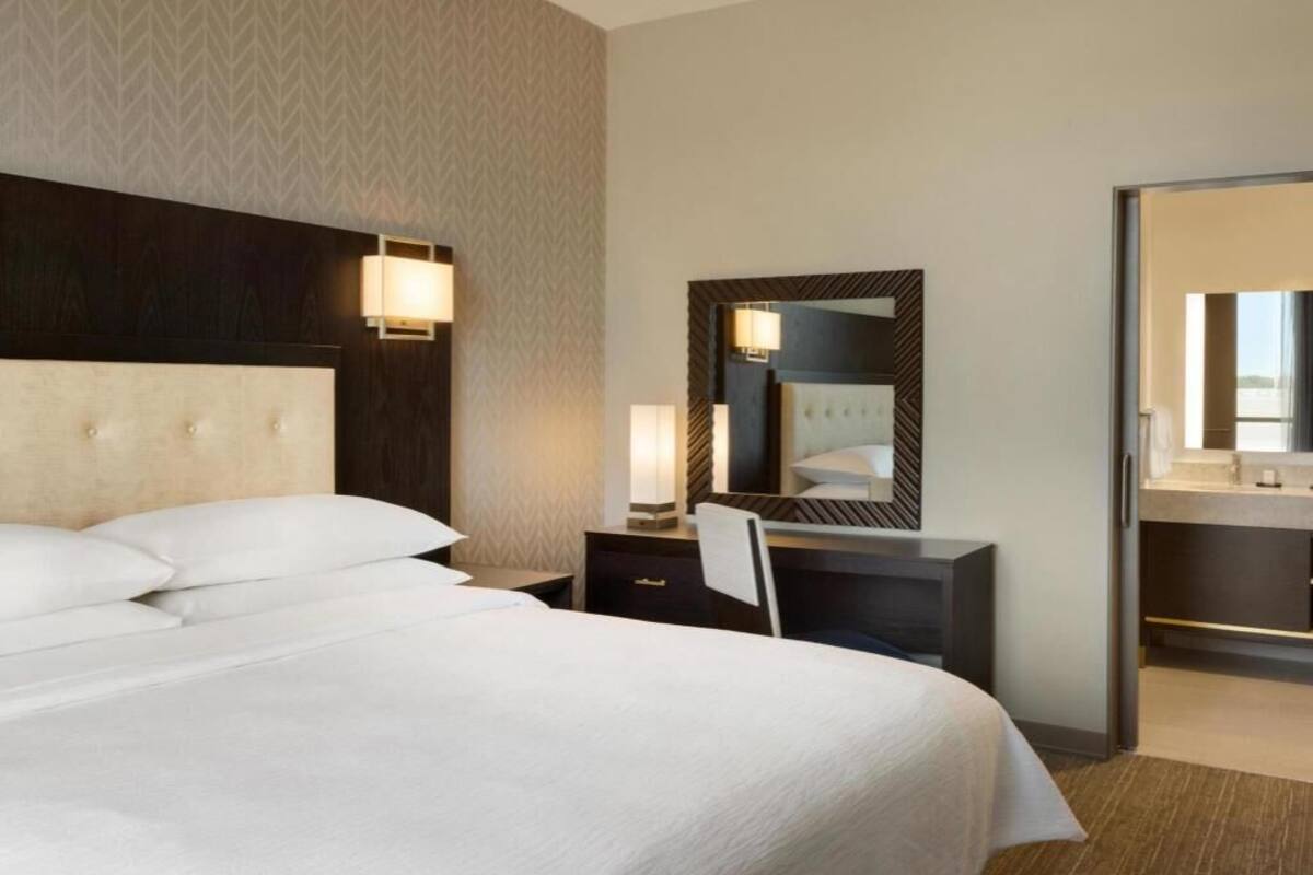 Een hotelkamer met een groot bed, een bureau met stoel, een spiegel en een badkamer die door een open deur zichtbaar is.