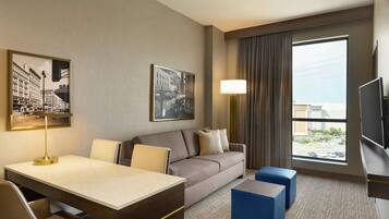Une chambre d’hôtel avec un canapé, un bureau avec une lampe, une télévision et une grande fenêtre donnant sur la ville.