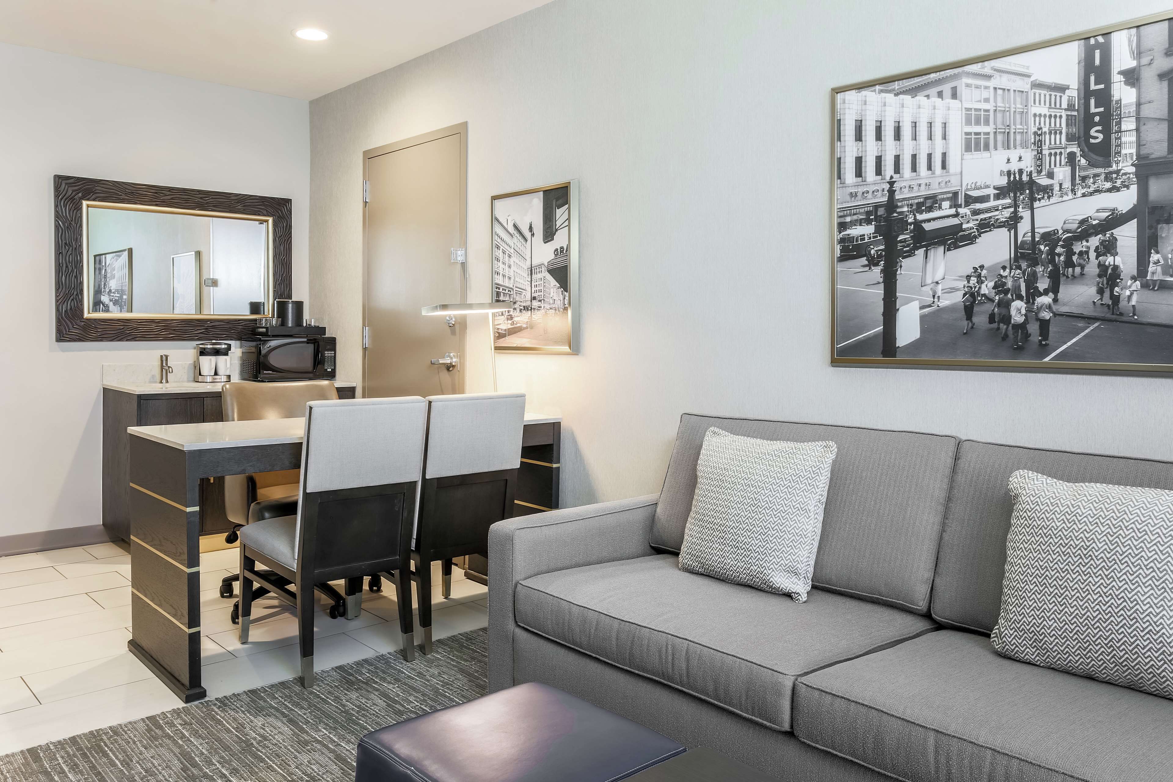 Habitación de hotel moderna con un sofá gris, un escritorio con una silla, un microondas y una cafetera. Hay una fotografía blanco y negro enmarcada en la pared.