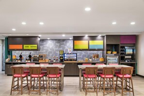 A modern restaurant with a central counter, multiple seating areas, and a variety of food and beverage stations. - Home2 Suites by Hilton Pensacola I-10 at North Davis Hwy (Pensacola)