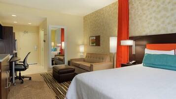 A hotel room with a bed, a desk with a computer, a chair, a sofa, and a nightstand with a lamp.