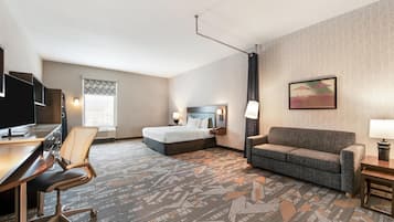 A hotel room with a bed, a sofa, a desk with a chair, a TV, and a patterned carpet.