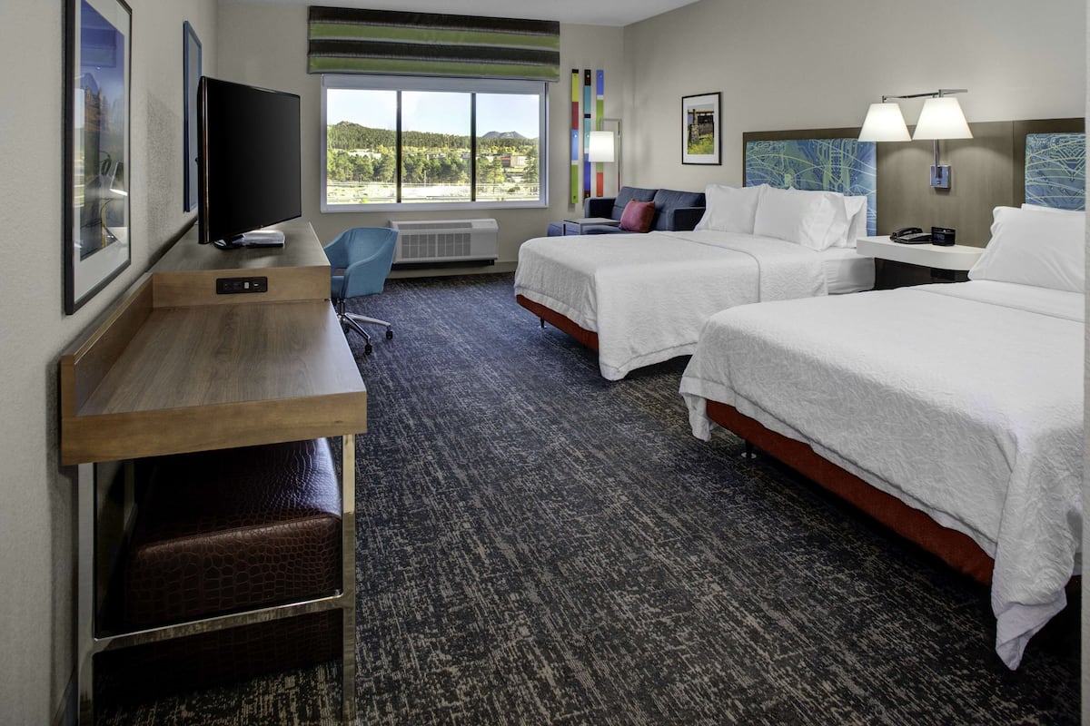 A hotel room with two beds, a desk with a flat-screen TV, a chair, and a large window offering a view of the outdoors.