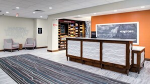 A hotel reception area with a reception desk, two armchairs, a patterned rug, a bookshelf with snacks, and a large 'explore.' poster on the wall. - Hampton Inn & Suites Flagstaff East (Flagstaff)