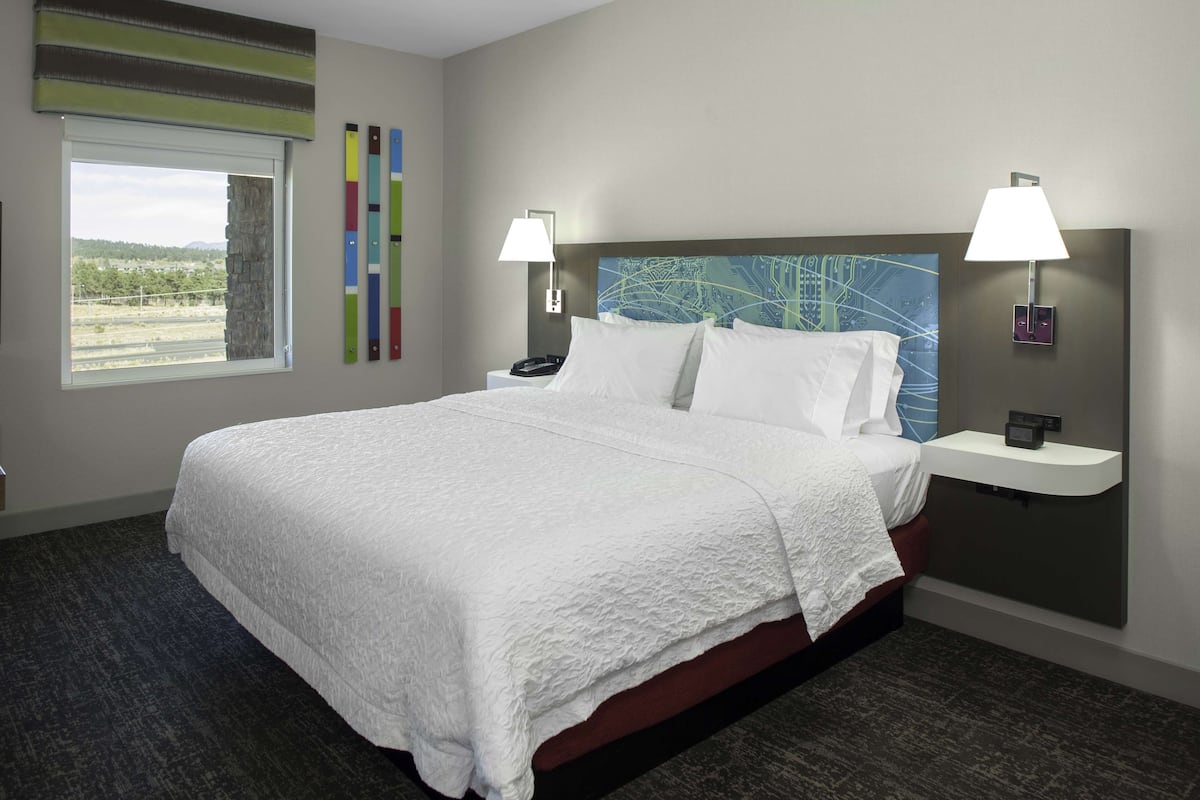 A hotel room with a large bed, two bedside tables with lamps, a window with a view of the countryside, and a wall with abstract art.