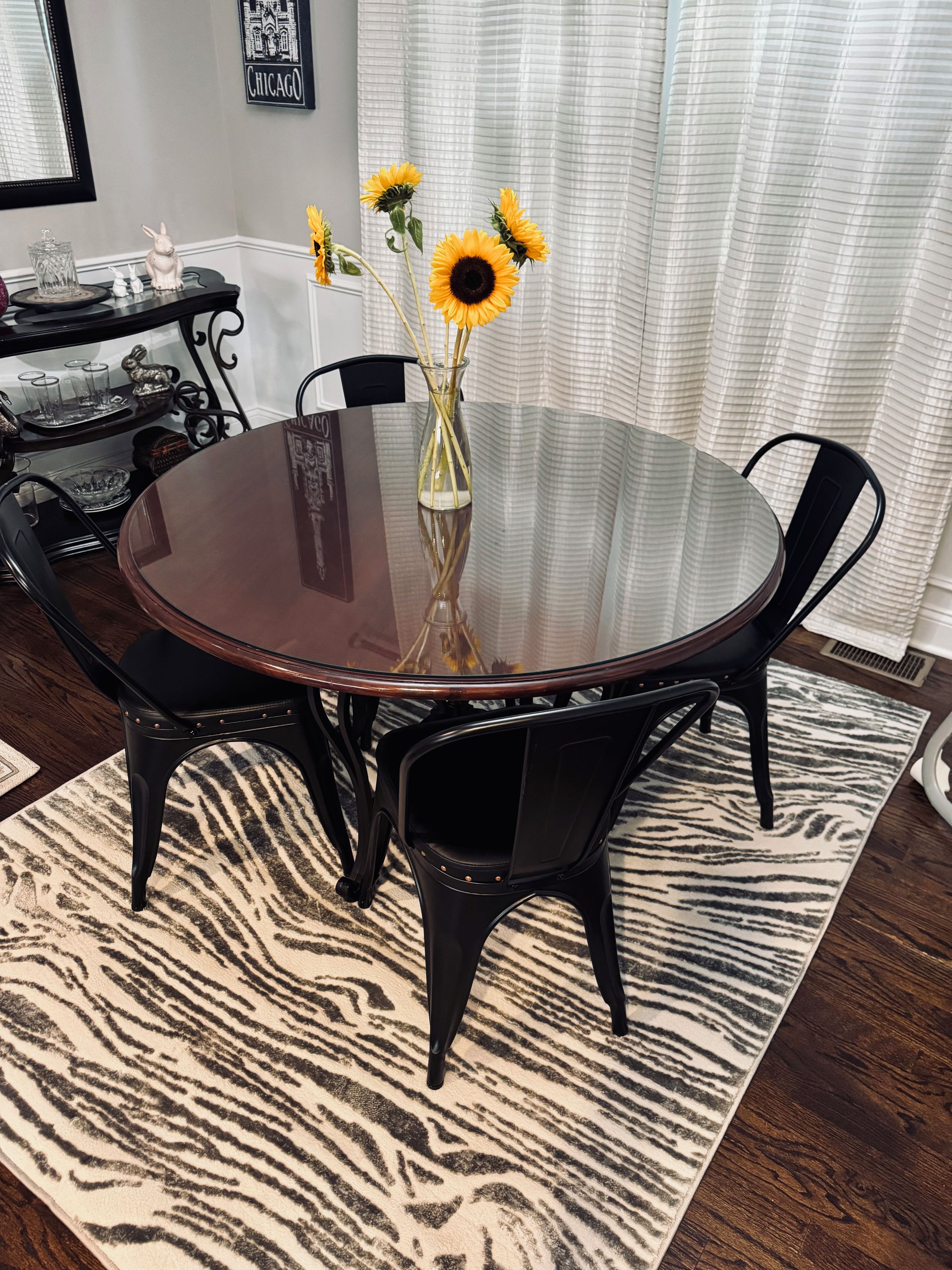 A round glass table with four chairs, a vase with sunflowers, and a zebra-patterned rug.