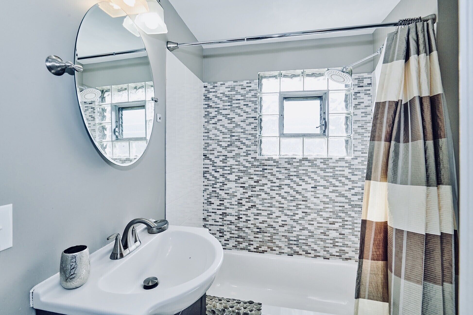 A bathroom with a white sink, a mirror, a shower with a tiled wall, and a window.