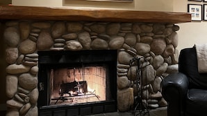 Una chimenea de piedra con repisa de madera, un cuadro abstracto enmarcado encima y un sillón de cuero negro a la derecha.