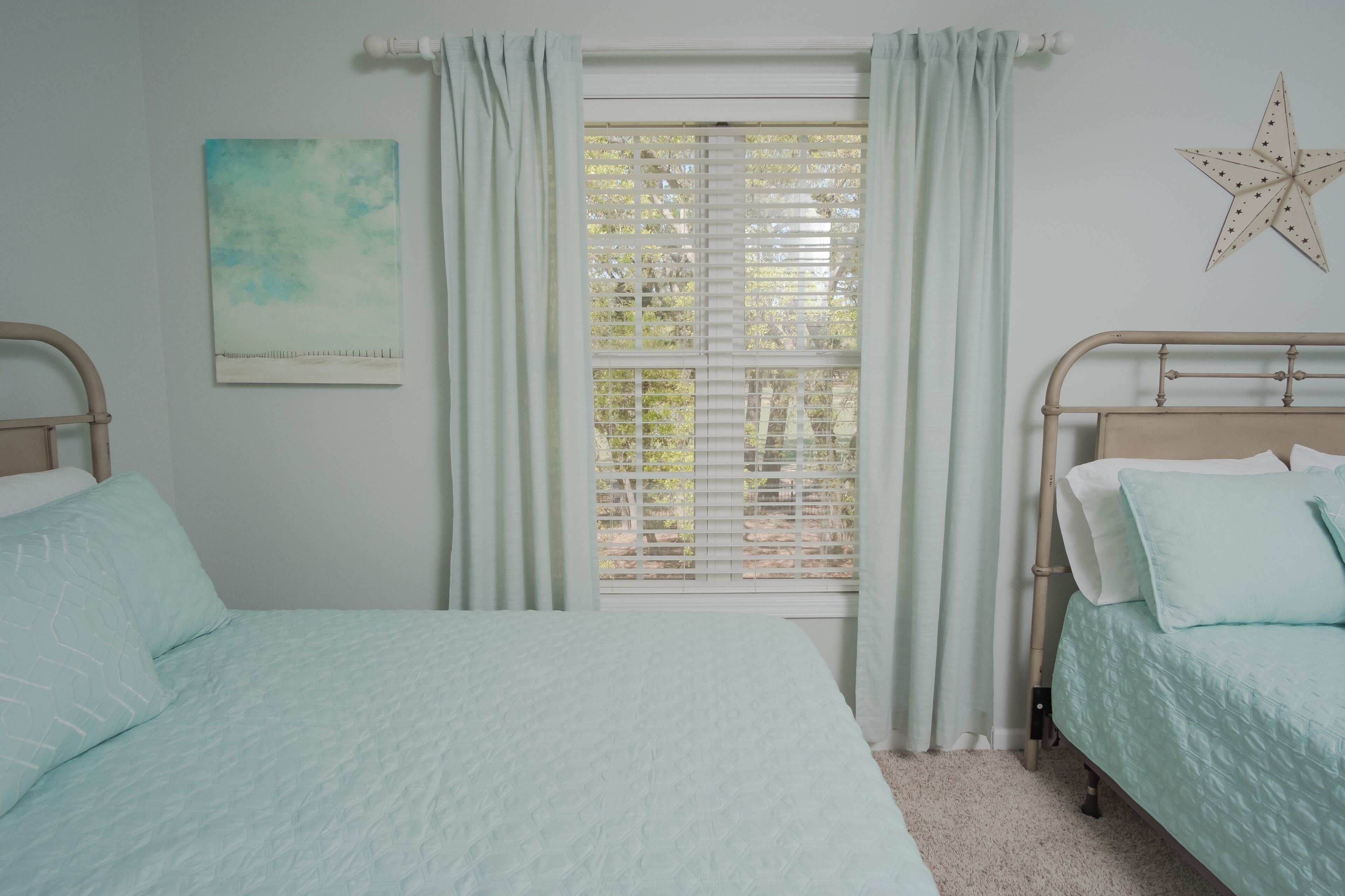 A bedroom with two beds, a window with blinds, a star decoration, and a painting on the wall.