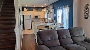 A modern living room with a dark sofa, a dining area with a table and chairs, and a kitchen in the background. - Beautiful Large Two Story Condo Near The Beach With Unique Pricing! (North Myrtle Beach)