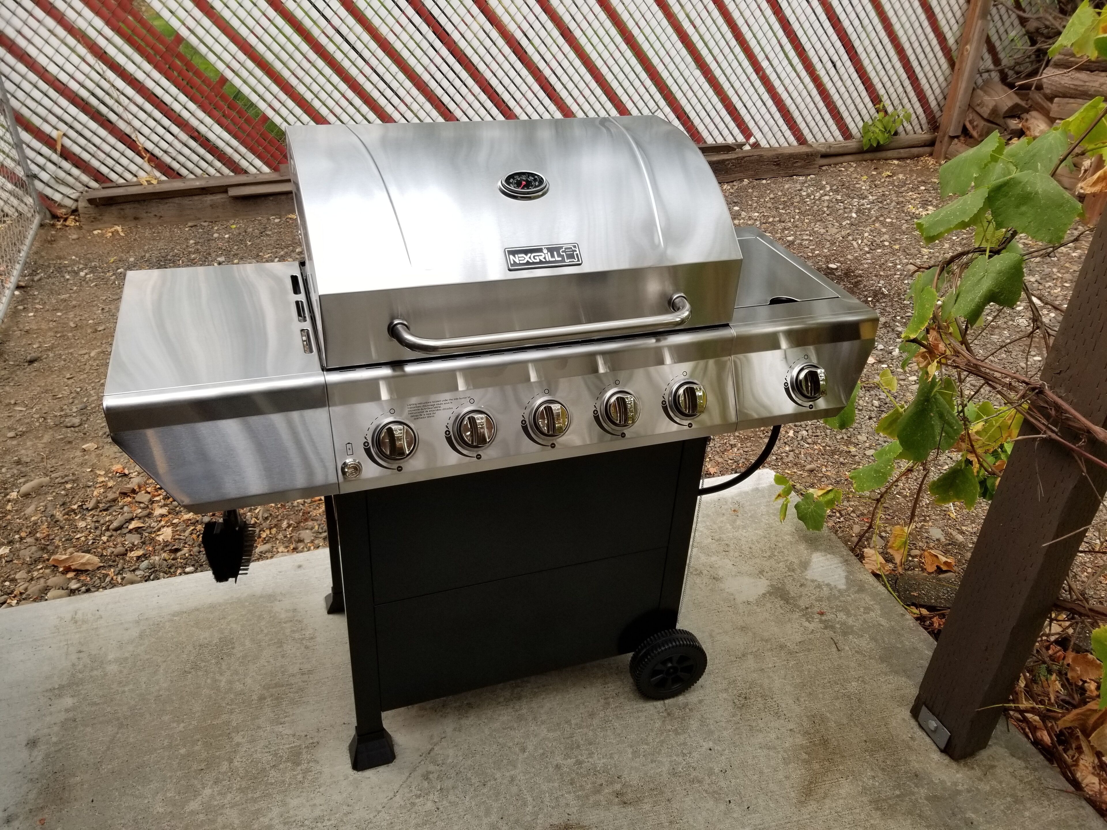 A stainless steel grill with four burners and a side shelf, placed outdoors on a concrete surface.