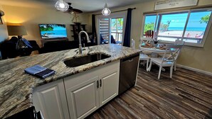 Una cocina con encimera de granito, pileta de acero inoxidable y muebles blancos. Hay una zona de comedor con mesa y sillas, y vistas a la playa a través de grandes ventanales.