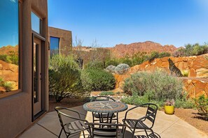 A patio with a round table and chairs, surrounded by desert vegetation and a mountain backdrop. - "SERENE DESERT GETAWAY" La Casa Coyote, Kayenta Art Village W/Views of Red Rock (Ivins)