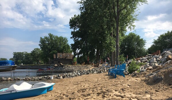 A beach with a row of blue chairs, a boat, and a rocky shoreline. - Lakefront, private beach, dock, kayaks, Paddle Boat and pet friendly! (Plattsburgh)