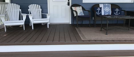 A porch with two white Adirondack chairs, a wicker chair with a cushion, and a small table.