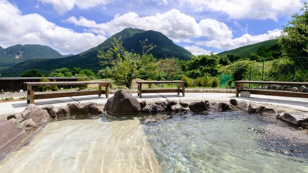A clear outdoor pool with wooden benches, surrounded by rocks and greenery, with mountains in the background. - Yufuin Tsukahara Villa ERINA (Yufu)