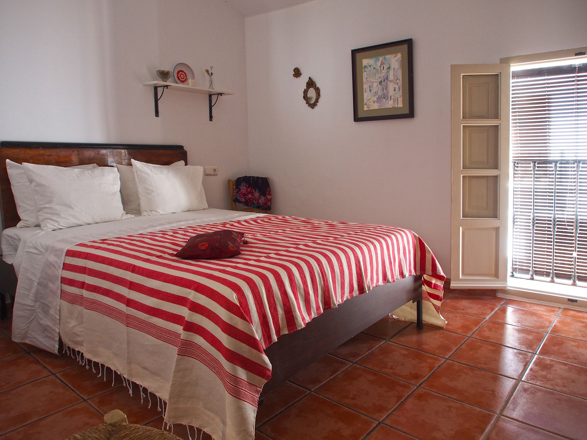 Une chambre à coucher avec un lit, des oreillers, une couvre-lit à rayures rouges et blanches, une petite étagère avec des objets décoratifs, un tableau encadré et une fenêtre avec des stores.
