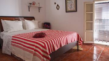 Une chambre à coucher avec un lit, des oreillers, une couvre-lit à rayures rouges et blanches, une petite étagère avec des objets décoratifs, un tableau encadré et une fenêtre avec des stores.