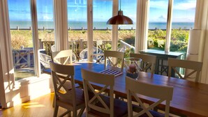 A dining area with a wooden table and chairs, a hanging pendant light, and large windows offering a view of the ocean. - Sea Front Luxury Beach House, Panoramic Views, Direct Beach access, West Sussex (Selsey)