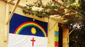 Eine Gebäudefassade mit einer Flagge, die ein Regenbogen, eine Sonne und ein Kreuz zeigt.