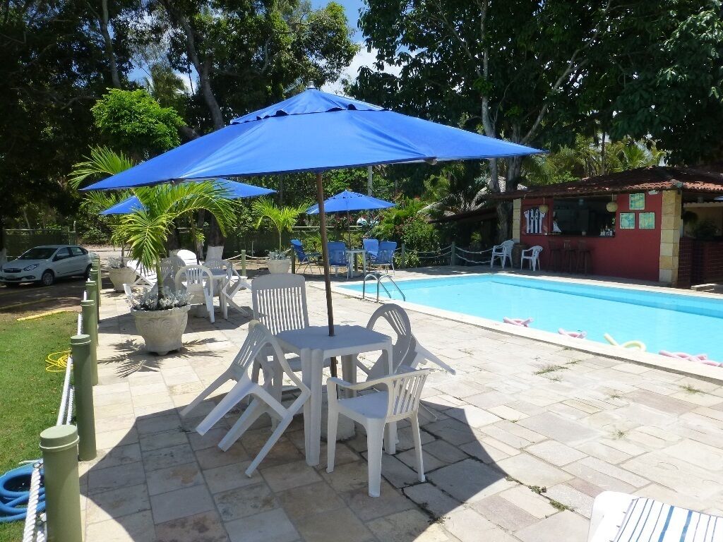 A poolside area with white plastic chairs and tables, blue umbrellas, and a swimming pool.