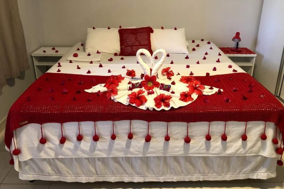 A neatly made bed with a red and white blanket, a heart-shaped decoration, and red flowers.