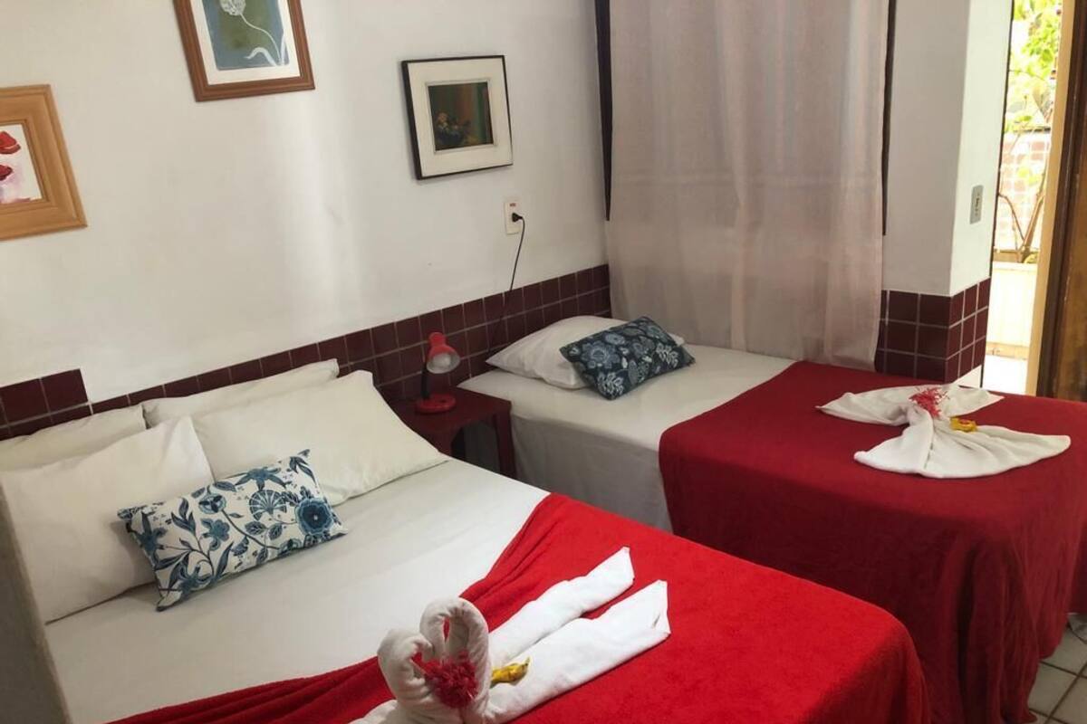 A hotel room with two beds, each with a red bedspread and white towels. There are framed pictures on the wall and a window with curtains.