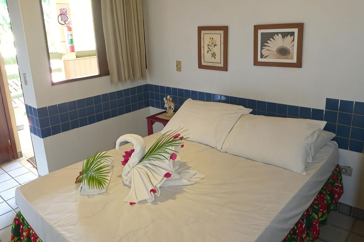 A hotel room with a bed, a window with curtains, a small statue on a nightstand, and two framed pictures on the wall.
