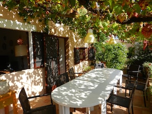 An outdoor dining area with a table and chairs, surrounded by greenery and a pergola with hanging lights.