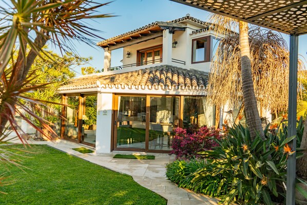 A two-story house with a patio, a covered outdoor seating area, and a garden with various plants. - RENOVATED 2026. BEACHFRONT. 5 MASTER SUITES. HEATED POOL. GAMES ROOM. (Marbella)
