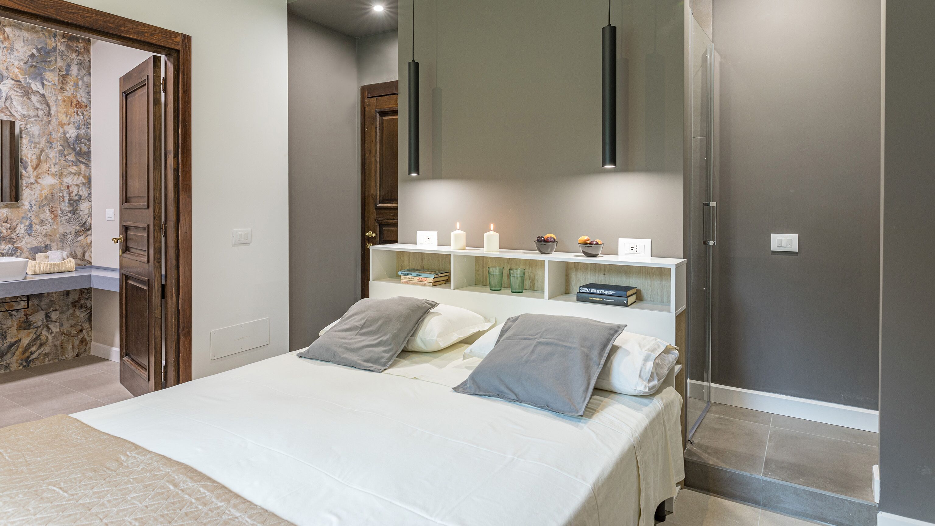 Quarto com cama, mesas de cabeceira, espelho e uma estante com livros e velas.