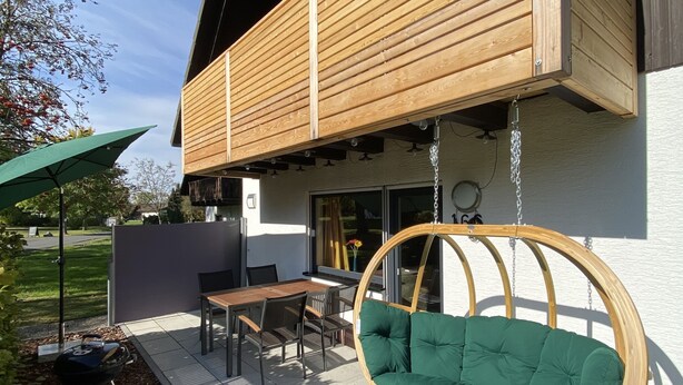 A modern outdoor patio with a wooden swing chair, a dining table with chairs, and a barbecue grill. - Beautiful vacation home on the Silbersee with extensive facilities and WLAN (Frielendorf)