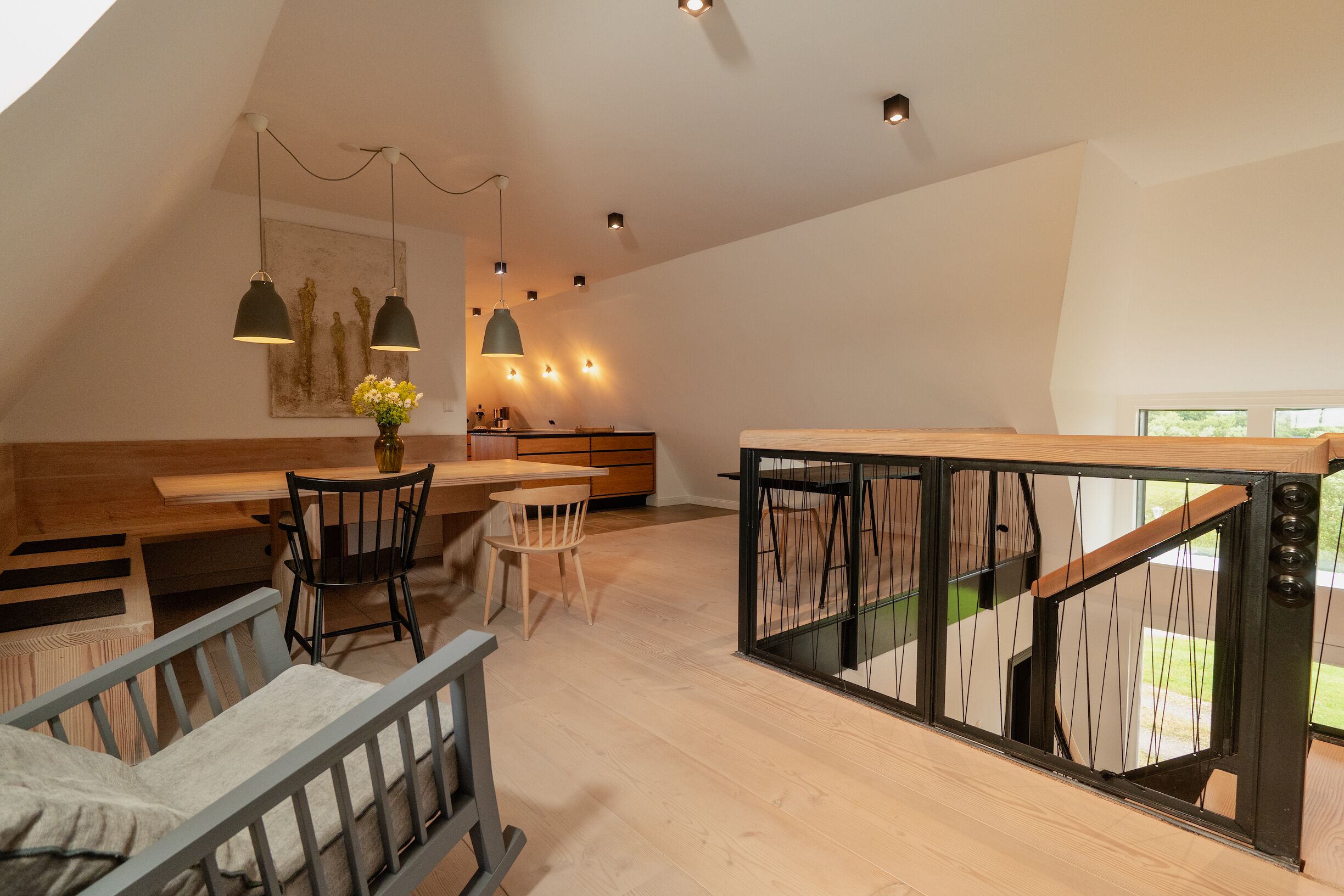 Een moderne loft-achtige kamer met een houten eettafel, stoelen, een bank en een trap met glazen balustrades.