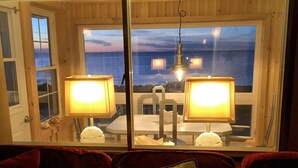 Une pièce chaleureuse avec un décor intérieur en bois, une fenêtre donnant sur la mer, deux lampes sur une table et un coussin orné d’un motif d’ancre.