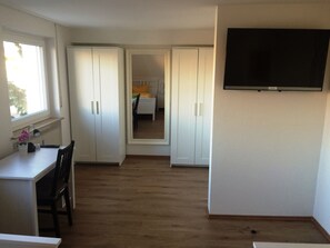 Une pièce de taille modeste comprenant un bureau, une chaise et un lit avec une table de chevet. On y trouve également une armoire à deux portes et un téléviseur fixé au mur.