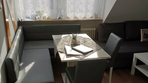 Ett mysigt rum med ett matbord, en bänk och en soffa. Det finns två fönster med gardiner, ett element och en hylla med dekorativa föremål.