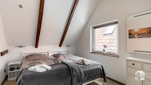 A bedroom with a sloped ceiling, a bed with a gray blanket, a window with a view of a building, and a small plant on the windowsill. - At the gates of Hamburg in the old country (Steinkirchen)