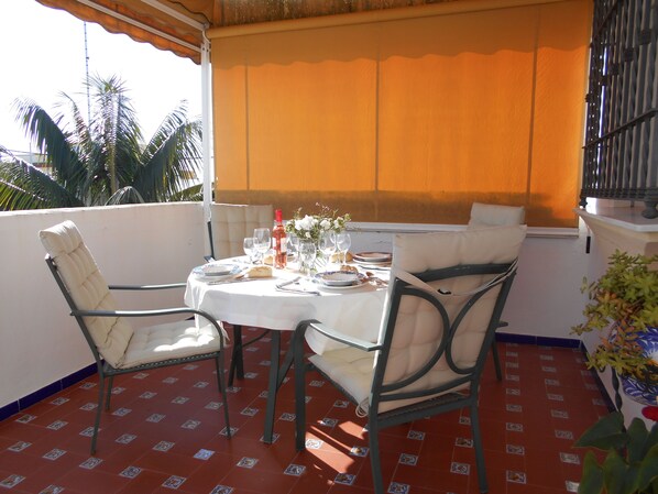 A round table with chairs, a bottle, glasses, and plates on a terrace with a tiled floor and a view of palm trees.
