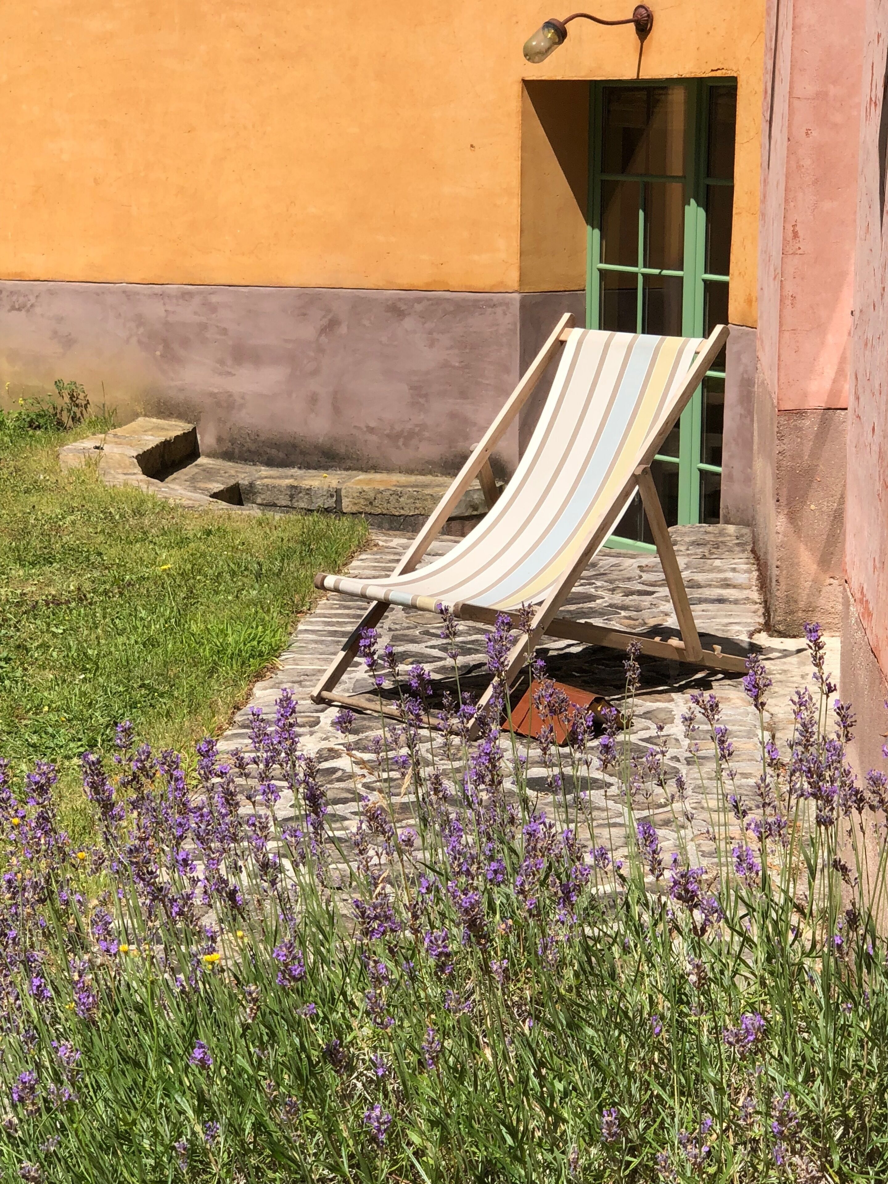 A striped lounge chair outside a building with a green door, surrounded by purple flowers and a stone pathway.