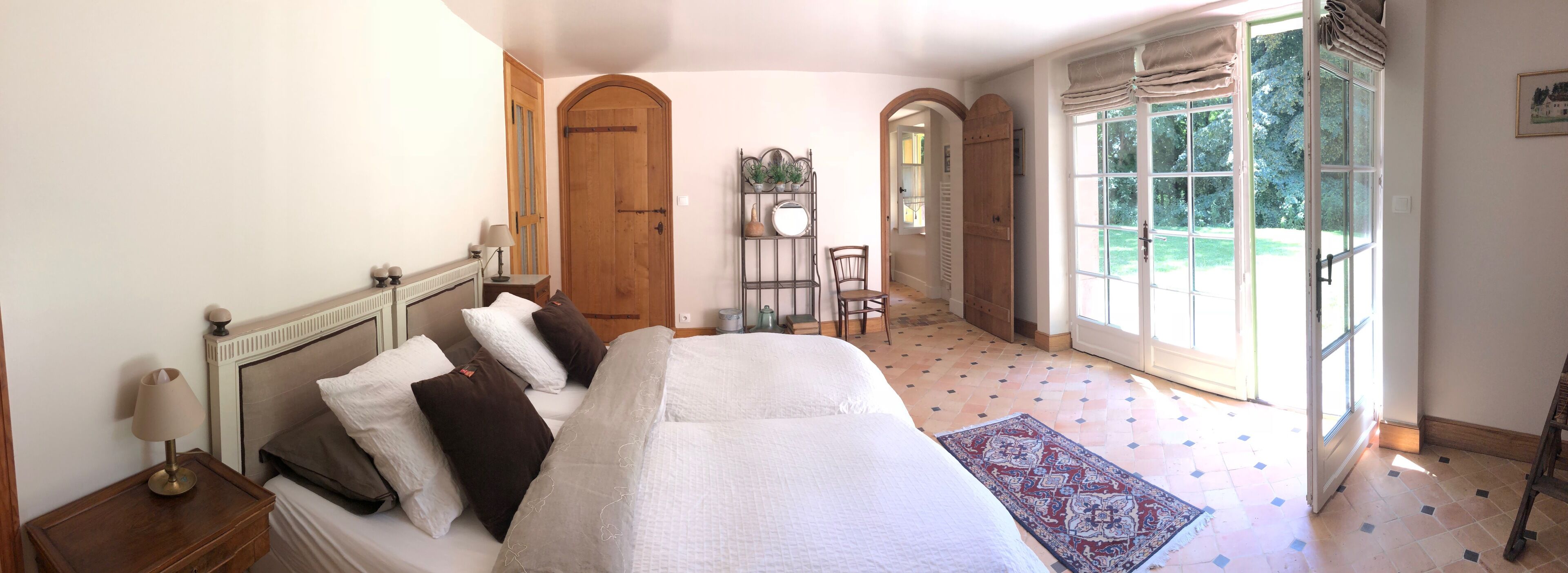 A bedroom with a large bed, a bedside table, a lamp, a chair, a window with curtains, and a door.