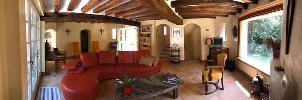 A spacious living room with a red sectional sofa, a glass coffee table, and a wooden chair. There is a bookshelf filled with books and a television on a wooden stand. The room has exposed wooden beams on the ceiling and large windows with a view of greenery outside. - Castle in the woods with private SPA 45 min from Paris and Reims, 25 min Disney (Montreuil-Aux-Lions)