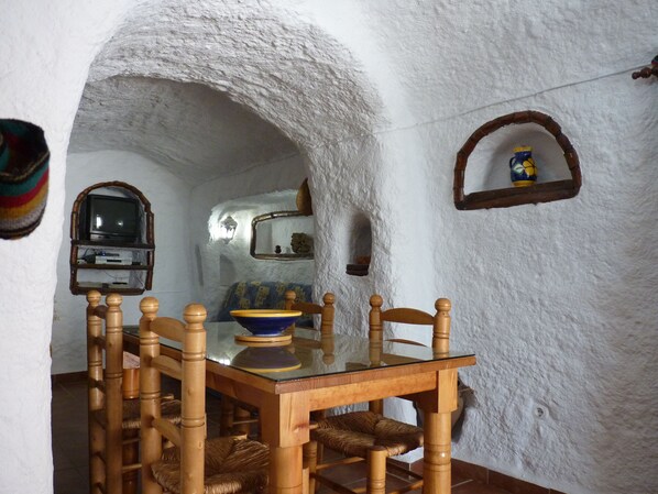 Un comedor rústico con una mesa y sillas de madera, un televisor en un estante y una pared blanca con textura.