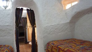 Una habitación con techo alto y blanco, con textura, una ventana pequeña y una puerta que da a otra estancia. Hay dos camas con ropa de cama de rayas coloridas.