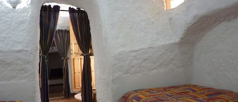 Una habitación con techo alto y blanco, con textura, una ventana pequeña y una puerta que da a otra estancia. Hay dos camas con ropa de cama de rayas coloridas.
