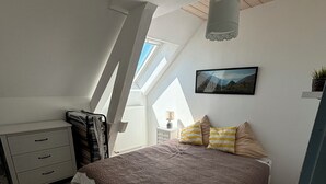 Une chambre avec un plafond en pente, un lit recouvert d’une couverture marron, une table de chevet avec une lampe et une fenêtre donnant sur l’extérieur.