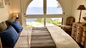 A bedroom with a large arched window, a bed with a white bedspread, blue pillows, and a wooden dresser with a lamp.