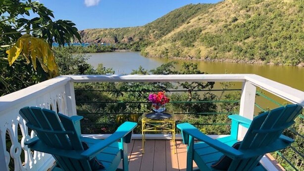 Eine Terrasse mit zwei türkisfarbenen Adirondack-Stühlen und einem kleinen Tisch mit Blumen, mit Blick auf einen See und Hügel.