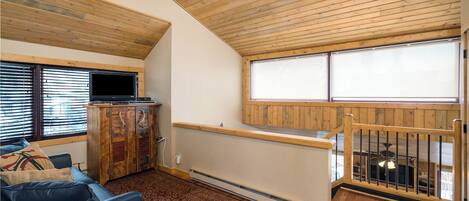 A cozy room with a wooden ceiling, a television, a cabinet, a sofa, and a window with blinds.